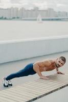 Flexible fit man does plank exercise on promenade has naked torso concentrated away prepares for workout practices yoga dressed in sportswear poses outdoor trains in morning. Sporty lifestyle photo