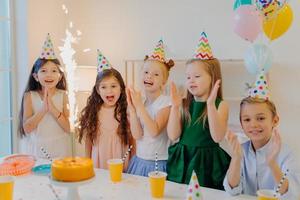 Cheeful friends of five pose near festive table with big cake, cup of drinks, wear party hats, look joyfully at sparkle, laugh happily, pose indoor, celebrate birthday
