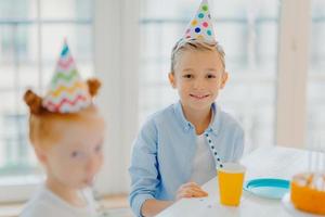 Happy small boy wears party hat, sits at table near ginger girl, have party together, celebrate birthday, pose in big spacious room with window. Children and holiday concept photo