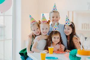 Five friendly little children wear festive cone caps, hug and make photo together, play games and celebrate birthday, have glad expressions, pose at festive table