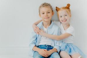 Friendly small girl and boy hug and sit on floor against white background, have positive expressions, feel satisfied after playing together, copy space. Ginger female kid embraces brother with love photo