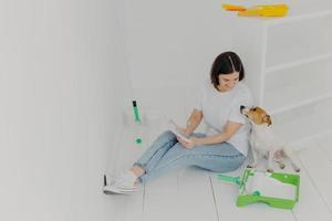 Horizontal shot of lovely woman with short hair, looks attentively at color palette, chooses colour for painting room, plays with pedigree dog, sit in white spacious room, sits on white floor photo