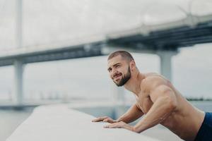 Strong bearded Caucasian man trains muscles and does push ups exercises, tries to reach fitness goals, demonstrates his physical strength, has naked torso, poses outdoor over bridge background. photo