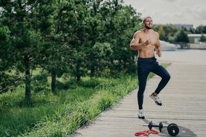 Horizontal shot of naked active man with naked torso jumps on one place, does sport exercises, poses outdoor, wears sport trousers and sneakers, warms up before jogging. Active lifestyle concept photo