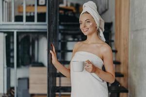 Photo of pleased smiling European woman enjoys day off at home, applies face cream, drinks tea, wrapped in towel, poses near strairs, thinks about something pleasant, has healthy glowing skin