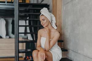 Thoughtful young European woman keeps hand under chin, sits on stairs and has thoughtful expression, has healthy skin, puts face cream, wrapped in bath towel, undergoes beauty treatments at home photo