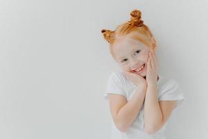 Happy small redhead girl tilts head, keeps hands on cheeks, smiles gently at camera, has good mood, rejoices spending free time with friends, dressed casually, isolated over white background photo