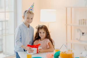 Handsome small boy gives present box to girl, celebrate birthday together, wear festive clothes and party cone hats, pose at table with cake in white spacious room. Holiday concept photo