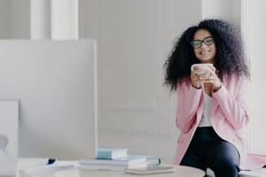 Joyful curly haired female freelancer or blogger has coffee break, holds white mug with beverage, poses near window, focused in computer, watches useful training video, wears near formal clothes photo