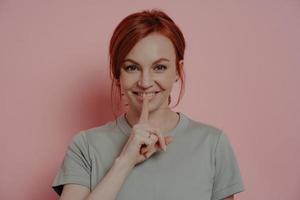 Young beautiful smiling red-haired woman telling secret information, posing isolated over pink wall photo