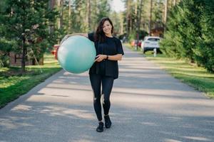 Young fit brunette woman holds big fitness ball, dressed in active wear, does exercises workout, poses in park, walks outdoor and breathes fresh air. People, gymnastics and aerobics concept. photo