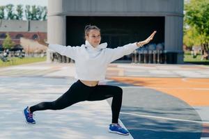 Active young woman dedicated to fitness does stretching exercises spreads hands dressed in active wear stretches legs and arms poses outdoo. People regular sport activities and lifestyle concept photo