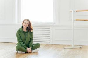 Photo of satisfied foxy European woman keeps legs crossed, poses near ballet barre, takes rest after dancing, wears green tracksuit, white sportshoes, stretches herself, models in dancing studio