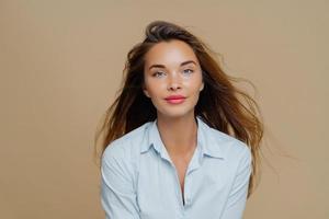 Lovely young female has long wavy hair floating in wind, dressed in elegant blue shirt, wears lipstick and makeup, poses against brown background, has direct gaze at camera, healthy facial skin photo