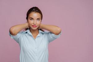 Indoor shot of brunette woman presses hair, has makeup, well cared complexion and fresh healthy skin, dressed in shirt, going to have walk with boyfriend, isolated over purple wall, copy space aside photo