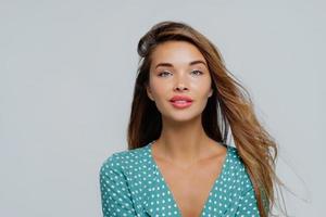 retrato de mujer joven de aspecto agradable mira tranquilamente a la cámara, tiene el pelo largo, vestido con una blusa de lunares, posa sobre fondo blanco, tiene belleza natural, tiene la cara y el maquillaje bien cuidados foto