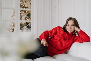 Indoor shot of pretty dark haired woman in warm winter clothes, leans at hand, lies on comfortable white sofa, looks directly at camera with calm expression. Domestic atmosphere. Time at home photo