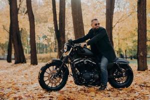 Thoughtful biker rides fast motorcycle, turns away, notices something into distance, wears sunglasses, poses in autumnal park, drives in nature. Lonely motorcyclist poses outdoor at nature during trip photo