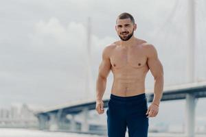 Confident cheerful bearded man with naked torso looks gladfully at camera, dressed in trousers, poses against river bridge, leads healthy lifestyle, has muscular body after regular trainings photo