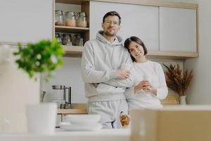 Indoor shot of pleasant looking wife leans at shoulder of husband, hold takeaway coffee, pose in kitchen, jack russell terrier dog poses near, cardboard boxes with personal stuff, modern interior photo