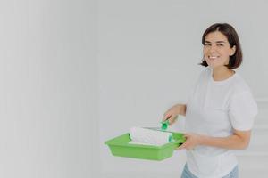 una mujer feliz y encantadora con una camiseta casual blanca, posa con un rodillo de pintura y una bandeja especial, hace reformas en un apartamento nuevo, tiene el pelo corto y oscuro y una sonrisa con dientes, aislada sobre fondo blanco. foto