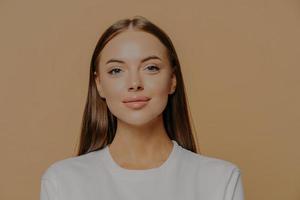 Portrait of pretty European woman with long hair looks confidently ar camera has healthy glowing skin makeup wears casual jumper isolated over brown studio wall. Beauty and wellness concept. photo