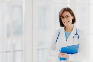 Good looking female expert creats medical prescription, writes down information in clipboard, thinks about good advice for patient, stands indoor, copy space for your information. Medicine concept photo