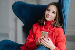 Pretty female teenager enjoys free time and popular music in earphones, holds modern smart phone, dressed in red knitted sweater, sits in comfortable armchair, uses application, checks new updates photo