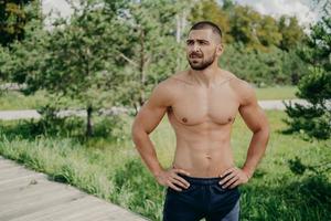 Horizontal shot of thoughtful shirtless sportsman keeps hands on waist, looks away demonstrates his muscular body, poses over nature background. Active bodybuilder goes in for sport, has daily workout photo