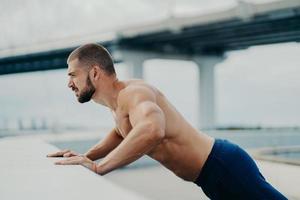 Horizontal shot of fit man with strong arms muscular body does push up exercises outdoor, has thick beard, concentrated into distance, goes in for sport regularly. Fitness and sporty lifestyle concept photo