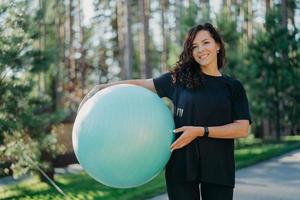 Positive brunette woman leads active lifestyle holds fitness ball poses against forest background dressed in black clothes has happy smile. Pretty female prepares for workout pilates does exercises photo