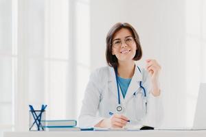 Young woman doctor writes on paper in clinic, creats prescription for patient, does paperwork in hospital, wears medical white gown, sits at cozy modern office. People, occupation, job concept photo