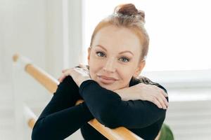 Beautiful young red haired female dancer crosses hands and leans on ballet barre which supports her during exercises practices dance in studio does stretching, being flexible looks directly at camera. photo