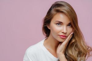 Close up shot of self confident beautiful Caucasian woman has long hair combed on one side, touches cheek with palm, wears minimal professional makeup, looks at herself in mirror, prepares for date photo