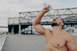 Tired sportsman cools off with fresh water poses with naked torso outdoor, tries to refresh himself, leads active lifestyle, takes break after cardio training. Excercising, refreshment concept photo