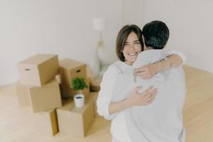 Indoor shot of lovely family couple embrace with love, hold keys from new apartment, move in own apartment, pose in empty light living room with floor lamp and pile of many cardboard containers photo