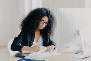 Photo of serious Afro woman writes in papers, sits at table with modern computer, creats article in newspaper, wears glasses and eyewear, poses indoor, works remotely. People and job concept