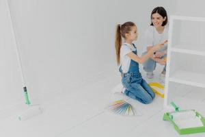 Busy girl and her smiling mother paint drawer in white color, use paintbrush or roller, surrounded with color samples, pose in white empty room, think about house decoration. Domestic work concept photo