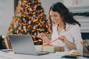Smiling woman unpacking Christmas gift while sitting at work desk with laptop against xmas tree photo