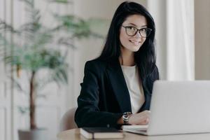 sonriente secretaria joven bien vestida con cabello oscuro, trabaja en una computadora portátil en el escritorio, hace proyectos o trabajos de investigación. gerente de ventas en oficina moderna, lugar de trabajo acogedor. concepto de trabajo remoto foto