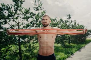 Horizontal shot of sporty man with bare chest exercises with elastic expander, gains for strong muscular body, has thick bristle, poses outside. Bodybuilder stretches band stands near green trees photo