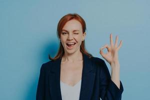 Young positive redhead woman in classic outfit showing okay gesture with hand and winking photo