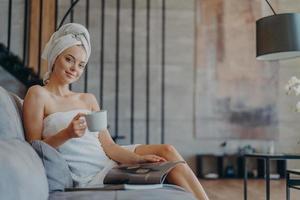 Pleased relaxed woman has rest at home poses on comfortable sofa wrapped in bath towel reads magazine drinks coffee poses over cosy home interior feels refreshed and pleased. People leisure. photo