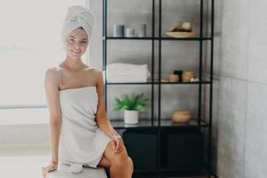 Pleased relaxed smiling woman with bath towel around healthy naked body, poses in bathroom with cosmetic product, takes care of herself, has skin care treatments. Spa, wellness, hygiene concept photo