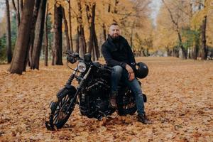 Bearded biker poses on own motorcycle, holds helmet, rides motorbike, poses outdoor in park, ground covered with fallen leaves. Biker lifestyle. Adventure on own transport. Male rider outdoor