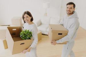 Positive female and male property owners pose with personal belongings in carton boxes, relocate to own apartment, smile pleasantly, pose in white living room. Moving day, planning, relocation photo