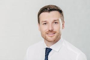 Headshot of attractive male entrepreneur with bristle, dressed in formal white shirt and tie, looks positively at camera, rejoces successful work in office, poses alone against white background photo