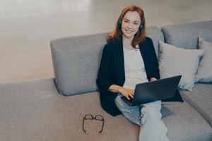 Beautiful smiling woman with red hair sitting with laptop and wireless headset on sofa in office photo