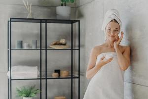 Horizontal shot of young healthy female model uses cream moisturizer, holds jar of cosmetic product, wrapped in towel after showering, stands near wall in cozy bathroom. Skin care procedures photo
