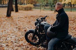 Back view of bearded male biker looks thoughtfully somewhere into distance, poses on black motorbike, holds protective helmet, spends leisure time in autumnal park, copy space for your advertising photo